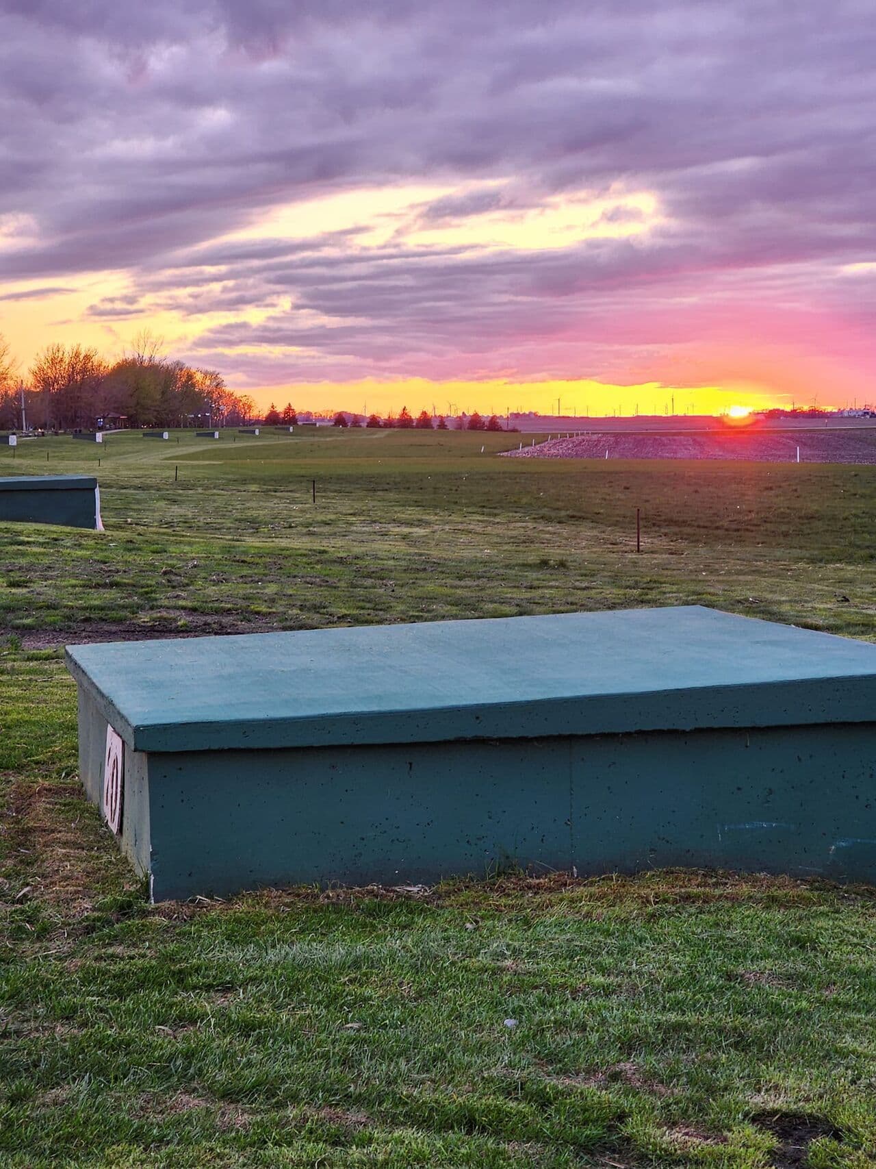 Stockdale Gun Club at sunset — teal trap house in foreground, cornfield horizon, wind turbines on the skyline